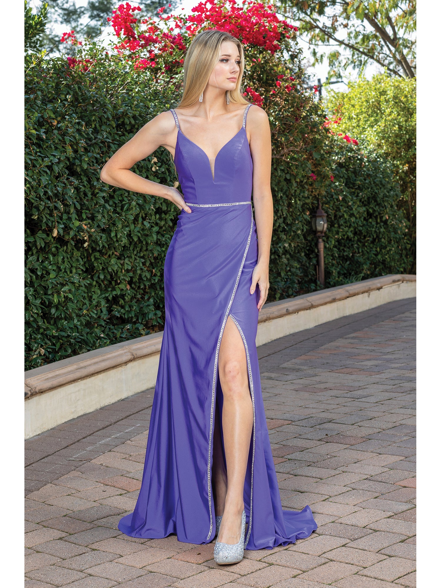 A woman stands on a paved walkway in front of green bushes and red flowers, wearing the Corset-Back Long Prom Dress with Beaded Accents, featuring a sleeveless design and a thigh-high slit.